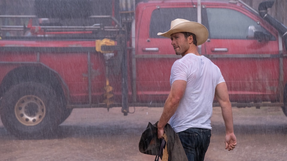 Tyler walks in the rain wearing a white t shirt and a cowboy hat