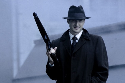 Sam Adama (Sasha Roiz) holds a gun while wearing a hat, glasses, and trench coat.