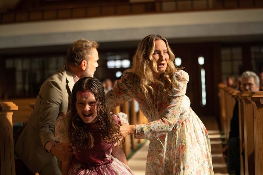 Possessed Katherine (Olivia O’Neill) is covered in blood in between pews of a church as Tony (Norbert Leo Butz) and Miranda (Jennifer Nettles) pull her back.