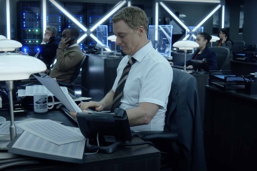 Harry Vanderspeigle (Alan Tudyk) works at a desk in Resident Alien Episode 301;