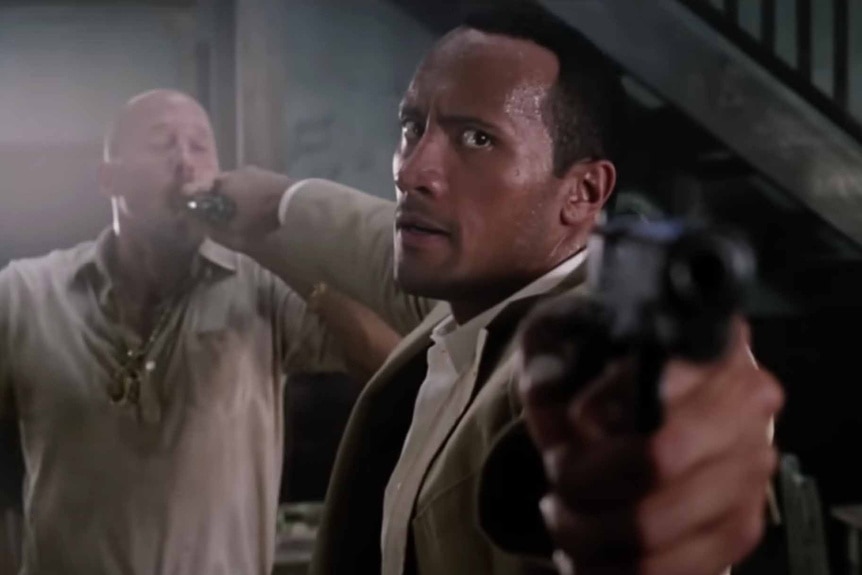 Beck (Dwayne Johnson) holds a gun in a man's mouth while pointing another in The Rundown (2003).