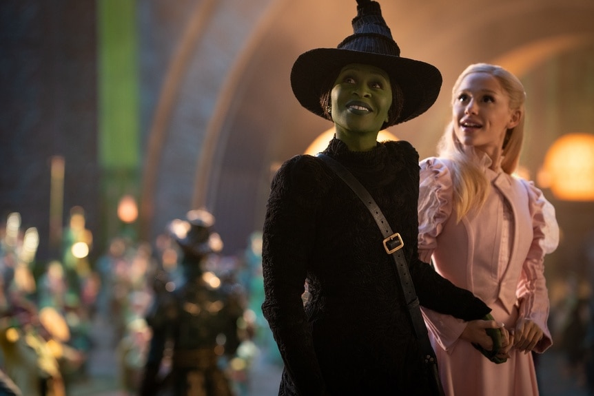 Elphaba (Cynthia Erivo) and Glinda (Ariana Grande) hold hands and smile in Wicked (2024).